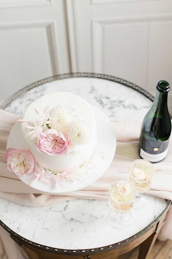 A close-up detail shot of a single-tier white fondant wedding cake decorated with fresh pink garden roses, white pompom dahlias, and small pink accent blooms, placed on a white cake board. The cake sits on a round marble-topped table with a silver filigree edge, styled with a draped blush pink silk fabric. Alongside the cake are two crystal champagne flutes filled with sparkling wine, each garnished with a small pink rose, and a dark green champagne bottle with a minimal label positioned to the right. The overall decor palette is white, blush pink, and soft ivory, with a refined, classic indoor styling approach against a white paneled wall background.