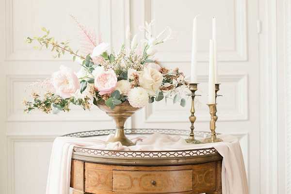 A close-up detail shot of a floral and decor arrangement placed on a small antique wooden side table draped with blush pink fabric. The centerpiece features a brass compote vase holding a loosely arranged bouquet of blush garden roses, ivory roses, eucalyptus, pink astilbe, dried grasses, and greenery. Two unlit white taper candles sit in brass candlesticks of varying heights to the right of the arrangement, all resting on a small mirrored or metal-rimmed tray. The backdrop is a white paneled wall, contributing to a classic French-interior aesthetic. The overall palette is blush, ivory, and antique gold with a romantic, vintage-inspired styling.