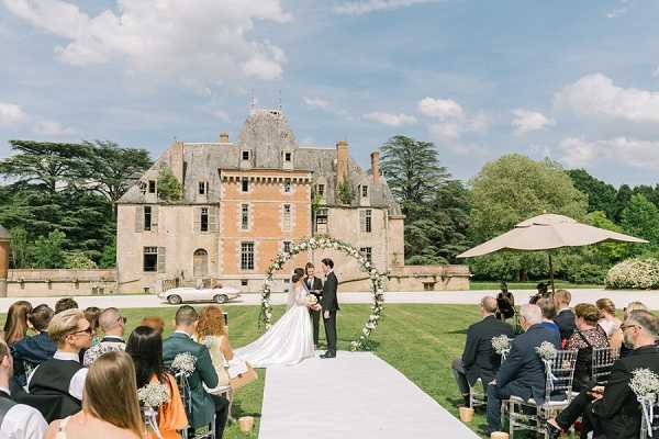 An outdoor wedding ceremony taking place on the grounds of a French chateau, with the couple standing beneath a circular floral arch decorated with white and green blooms at the end of a white aisle runner. The bride wears a long white gown with a train, and the groom is in a dark suit; they appear to be exchanging vows with an officiant present. Approximately 40–50 guests are seated on either side of the aisle in clear acrylic Chiavari-style chairs, with small clusters of white baby's breath visible on the chair ends as decoration. The ceremony setup has a classic, garden-party aesthetic with a white and green floral palette, set against the facade of a multi-story stone and brick French chateau with pointed turrets in the background. Wide shot taken from behind the seated guests, capturing the full scene including the venue architecture and manicured lawn. Potential venue feature image.