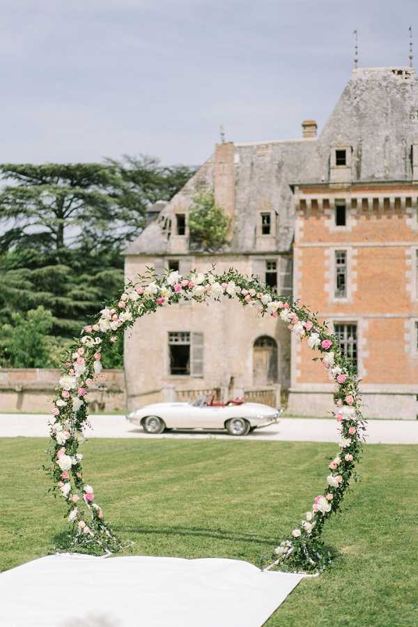 An outdoor ceremony setup on the lawn of a French chateau, featuring a full circular floral arch decorated with ivory and pink roses, blush blooms, and trailing green foliage. A white fabric aisle runner is laid on the grass in front of the arch. In the background, a white vintage convertible sports car — likely a Jaguar E-Type — is parked in front of the chateau, which features a mix of pale stone and red brick architecture with pointed slate rooftops typical of French Renaissance style. No people are present in the image. The composition is a wide portrait shot with the arch as the central foreground subject. Potential venue feature image.