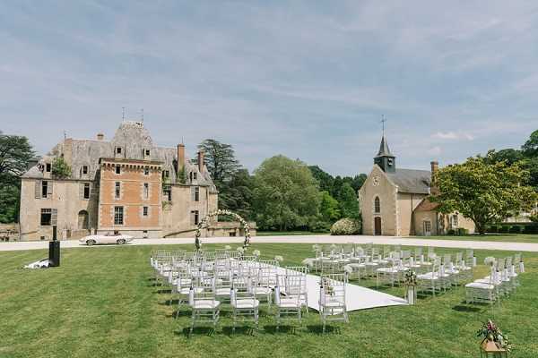 An outdoor wedding ceremony setup on the lawn of a French chateau estate, with a stone chapel visible to the right of the main castle building. White Chiavari chairs are arranged in a semicircular configuration around a white aisle runner, with a floral arch featuring white and greenery blooms at the altar. Small floral arrangements on stands mark the ends of the chair rows. The ceremony space is unoccupied, suggesting this is a pre-ceremony setup shot. The wide-angle composition captures both the chateau and chapel as backdrop elements alongside the full seating arrangement. Potential venue feature image.
