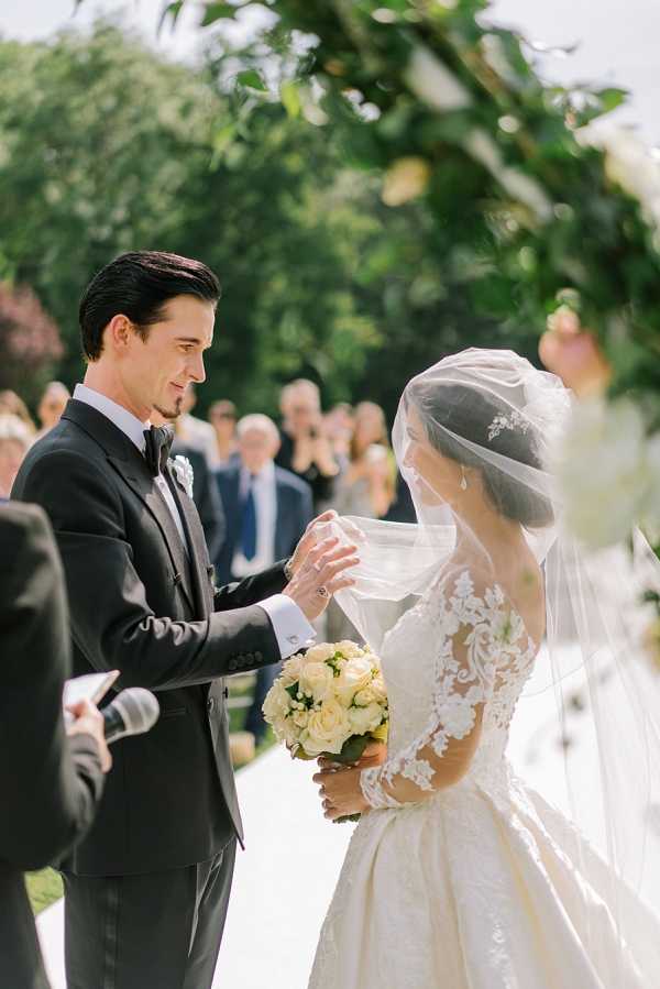 An outdoor wedding ceremony captured in a medium portrait shot, showing the groom lifting the bride's veil at the altar. The groom wears a dark charcoal tuxedo with a black bow tie and a white boutonniere, while the bride wears a full-skirted ivory ball gown with long lace sleeves and intricate floral lace detailing on the bodice, paired with a cathedral-length veil and a delicate hair piece. The bride holds a round bouquet of cream and ivory roses with greenery. A floral arch with lush greenery and soft blush and cream blooms frames the couple from above, and a row of seated guests is visible in the soft-focus background along a white aisle. The styling is classic and formal, with a refined, traditional aesthetic.