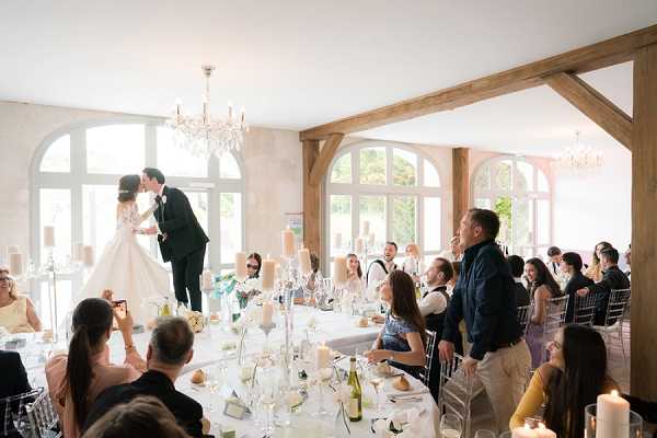 The bride and groom share a kiss while standing elevated at the head of a long reception dining table, with approximately 30 guests seated and standing around them watching and reacting. The indoor reception room features arched white-framed windows, exposed oak timber framing, and crystal chandeliers overhead. The bride wears a full-skirted white ballgown and the groom a black suit; the tables are dressed with white linens, tall pillar candles, white floral centerpieces, and glassware set for a formal dinner. The image is a wide shot capturing the full room and its light-filled interior with a classic, clean decor palette of white and ivory.