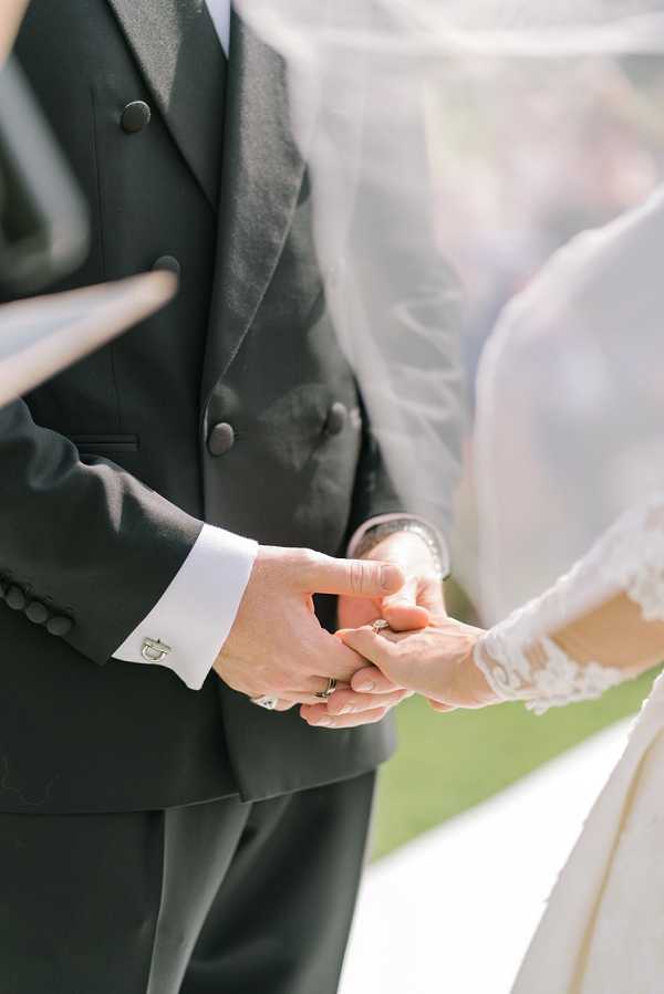 Close-up detail shot of a couple holding hands during what appears to be an outdoor ceremony. The groom wears a dark charcoal three-piece suit with a double-breasted waistcoat, white dress shirt, and silver cufflinks, with a wedding band visible on his finger. The bride wears a lace-sleeved white gown and a sheer veil that drapes across the right side of the frame, with a bracelet visible at her wrist. The image is tightly cropped at torso and hand level, with a softly blurred background.