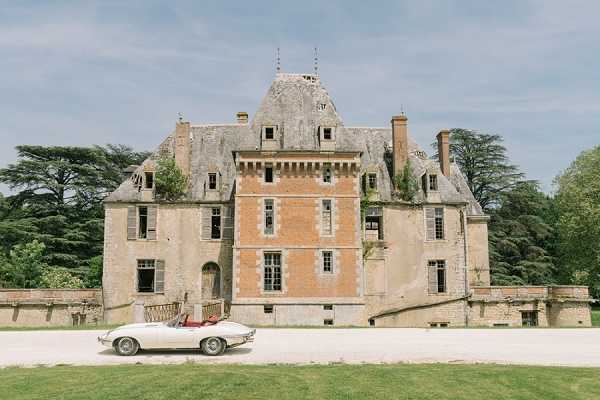 A wide exterior shot of a French chateau with warm terracotta and stone facade, steeply pitched slate roof with dormer windows, and symmetrical wings. A classic white convertible sports car with a red interior — likely a vintage Jaguar E-Type — is parked on the gravel driveway in front of the building, with what appears to be a figure seated inside. The composition frames the full chateau facade with the car as a focal point in the foreground, emphasizing the historic architecture. Potential venue feature image.
