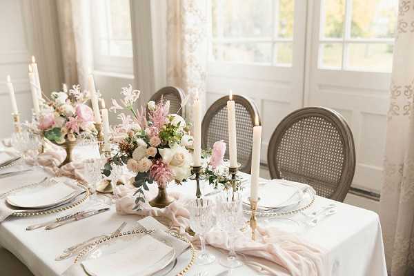A close-up detail shot of an indoor wedding reception tablescape styled in a soft, romantic palette. The table is set with a white linen, gold beaded charger plates, white dinnerware, crystal glassware, and silver flatware, with blush pink silk runners draped loosely across the center. Floral centerpieces in antique brass compotes feature ivory garden roses, blush pink astilbe, mauve blooms, and eucalyptus foliage. Tall ivory taper candles in brass candlesticks are arranged at varying heights along the table, and French cane-back chairs in a taupe finish are visible. The setting is in a bright interior room with large white-framed windows and sheer embroidered drapes, suggesting a chateau or manor house venue with a classic French-inspired aesthetic.