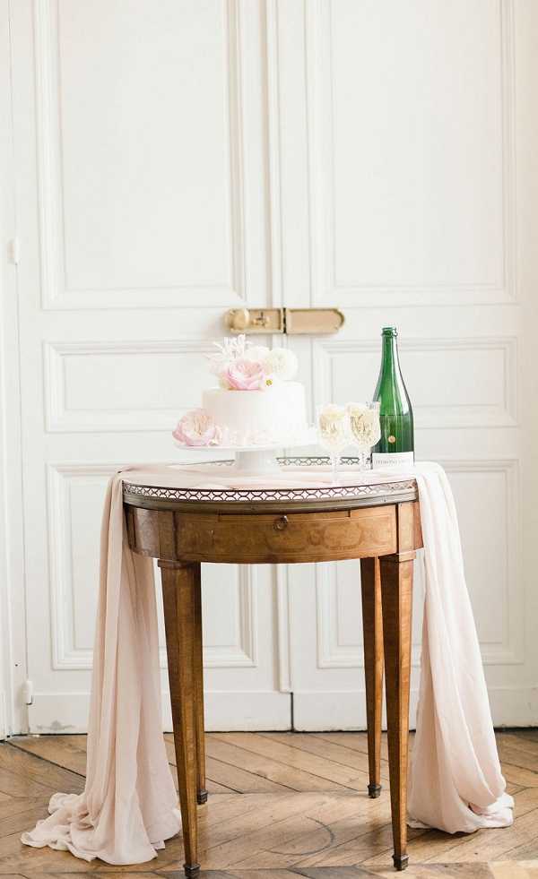 A detail shot of a wedding cake display set up indoors against white French boiserie paneling. A two-tier white fondant cake decorated with blush pink and ivory flowers, including what appear to be ranunculus and peonies, is placed on a white cake stand atop an antique walnut demi-lune table with tapered legs and a decorative brass gallery rail. Blush pink chiffon fabric drapes loosely over both sides of the table and pools onto the herringbone parquet floor. Two champagne flutes with gold monogram detailing and a green glass champagne bottle are positioned to the right of the cake. The overall styling palette is blush pink, ivory, and warm wood tones with a classic French interior aesthetic.