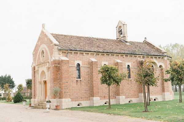 Wide exterior shot of a small red brick chapel with stone trim detailing, arched windows, a slate roof, and a small bell tower. The chapel features a wooden arched entrance door and is surrounded by neatly trimmed small trees and a gravel path. No people are visible in the frame. The image has a bright, airy exposure with an overcast white sky. Potential venue feature image.