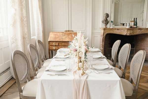 A styled reception tablescape shot indoors within a classic French interior featuring wood-panelled walls, a carved stone fireplace with a bust sculpture, and herringbone parquet flooring. The rectangular table seats approximately eight, dressed in a white linen tablecloth with a blush pink silk runner cascading down the center. Place settings include white plates, silver cutlery, and clear glassware, while a gold candelabra holds tapered ivory candles surrounded by a floral arrangement of blush pink roses, ivory dried pampas grass, and eucalyptus foliage. The chairs are grey-painted French Louis-style cane-back chairs, and an antique gilt writing bureau is visible in the background. The overall styling is classic French with romantic, soft blush and gold tones. Wide interior shot. Potential venue feature image.