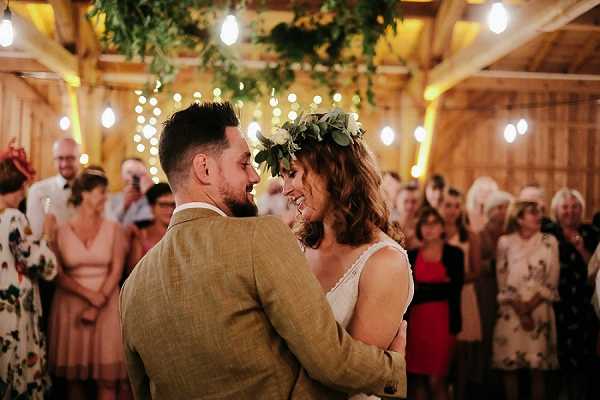 A couple shares their first dance inside a rustic barn venue, surrounded by approximately 20-30 guests standing in a circle to watch. The groom wears a tan/mustard tweed suit jacket and the bride wears a simple white dress with thin straps, accessorized with a green leaf crown on her loose wavy hair. The barn interior features warm amber uplighting on wooden walls, string Edison bulb fairy lights overhead, and a hanging greenery installation above the dance floor. The couple is framed in a close portrait shot from behind and to the side, with the smiling bride facing the camera. The styling is rustic-boho with a green and natural wood palette.