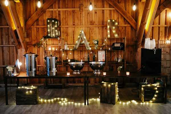 A wide shot of a rustic indoor bar setup at a wedding reception, set inside a wooden barn with exposed beams and warm wood-paneled walls. The bar consists of a long dark table holding two large silver punch bowls, a stainless steel coffee urn, glassware, and small candle votives. Behind the table, open wooden shelving displays bottles and small decorative items. Large marquee letters spelling 'BAR' with warm Edison bulbs are mounted on the back wall, and Edison bulb string lights are draped across the shelving and walls. Dark wooden crates in the foreground are wrapped with warm white fairy lights. The overall decor palette is dark and warm-toned, with a rustic style consistent with a barn wedding aesthetic.