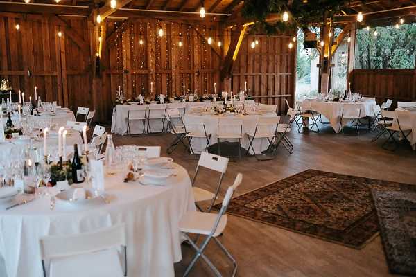 A wide shot of a wedding reception setup inside a rustic wooden barn venue. The space features a mix of round and long rectangular tables dressed in white linen tablecloths, set with glassware, wine bottles, and white taper candles as centerpieces with small low floral arrangements. Seating is provided by white and dark metal folding chairs. The barn's wooden ceiling and walls are decorated with strands of warm Edison bulb string lights and greenery garlands draped across the rafters. A patterned vintage-style area rug is placed on the wooden floor near the entrance area, adding a boho rustic touch to the overall decor scheme of white, warm wood tones, and green. No guests are present, indicating this is a pre-reception setup shot. Potential venue feature image.
