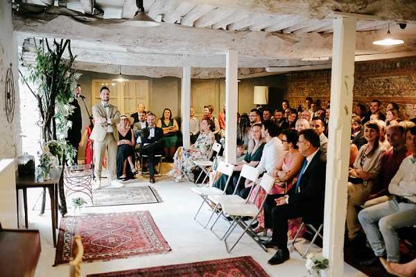 An indoor wedding ceremony is underway, with a man in a tan suit standing at the front speaking to a seated crowd of approximately 40–50 guests arranged in white folding chairs. The venue is a rustic converted space with exposed stone walls, whitewashed wooden ceiling beams, and white-painted structural columns. The ceremony area is decorated with red and burgundy patterned Persian-style rugs layered on a white floor, a tall greenery arrangement with trailing foliage, and a small side table holding a white floral arrangement. Guests are dressed in smart casual summer attire in a variety of colors. The shot is a wide-angle perspective taken from the back of the room, capturing the full depth of the space and the audience's attention directed toward the speaker.