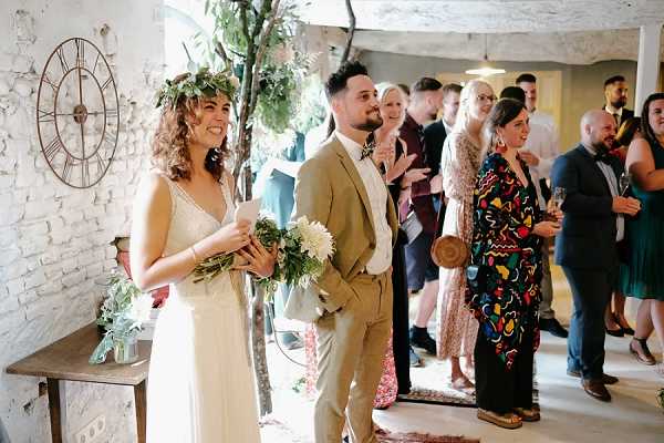 The bride and groom stand together indoors during what appears to be a reception or cocktail hour, surrounded by approximately 10–12 guests who are socializing and applauding. The bride wears a flowing ivory V-neck lace dress with a large green foliage crown and holds a loose bouquet of white flowers and greenery, reflecting a boho style. The groom wears a tan suit with a floral bow tie. The interior space features whitewashed brick walls, a large metal Roman numeral clock, a small wooden side table, and a potted tree, giving the venue a rustic, industrial character. The shot is a medium wide-angle candid capturing the couple's relaxed, joyful expressions alongside their guests.