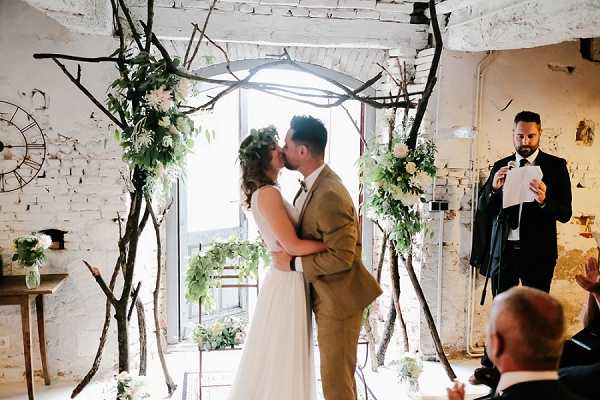 The bride and groom share their first kiss during an indoor ceremony in a whitewashed brick venue with an industrial aesthetic. They stand beneath a rustic arch made from natural bare branches, decorated with lush greenery and white florals, likely white roses or peonies. The bride wears a flowing white gown and a large greenery crown in her hair, while the groom is dressed in a tan suit with a bow tie. An officiant in a dark suit stands to the right reading from a paper, and at least one seated guest is visible in the foreground. The decor palette is white and green with a boho-rustic style, and small floral arrangements in glass vases are visible at the base of the arch and on a side table to the left. The shot is taken at mid-distance, capturing the full arch and the couple within the ceremony space.