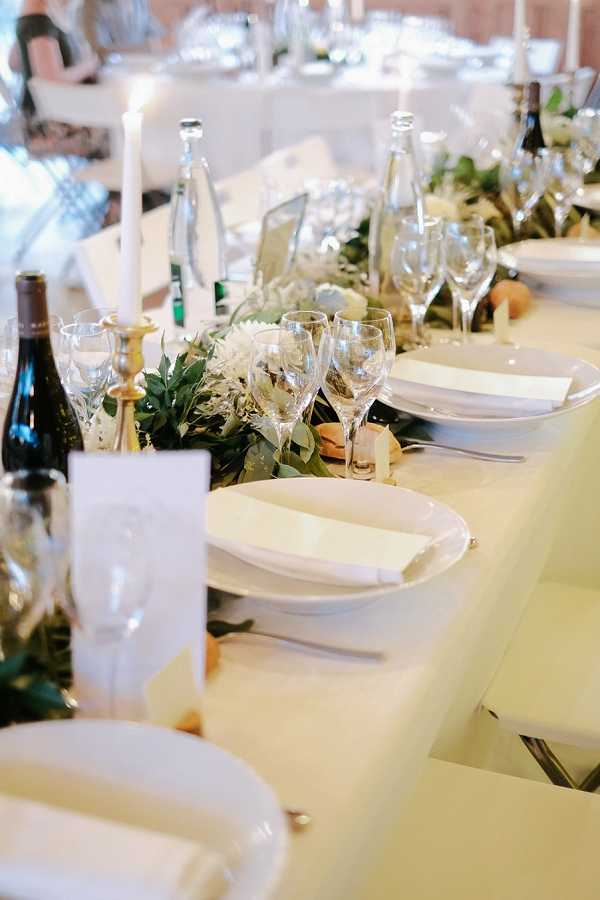 A close-up detail shot of a wedding reception table set for dinner, photographed from a low angle along the table's length. The table is dressed with a white linen cloth and features a lush greenery garland runner accented with white daisy-style blooms and what appear to be small fruits or citrus elements. Place settings include white round plates with folded white linen napkins, crystal wine glasses in multiple sizes, and silver cutlery. Tall white taper candles in gold candlestick holders are spaced along the center, alongside glass water bottles and wine bottles. Small white menu or place cards are visible at each setting. The overall decor palette is white, ivory, and green with gold accents, suggesting a classic, clean aesthetic. Guests are faintly visible and out of focus in the background within what appears to be an indoor reception venue with warm ambient lighting.