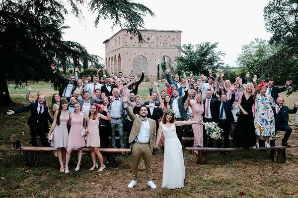 A large group wedding portrait taken outdoors, featuring the couple at the front center surrounded by approximately 50 guests, all with arms raised and celebrating. The groom wears a tan/camel suit with a bow tie and white sneakers, while the bride wears a simple white flowing gown and holds a loose bouquet with white flowers and greenery. Guests are dressed in a mix of smart-casual attire including blush pink bridesmaid dresses, dark suits, and colorful printed dresses. The setting is a garden or parkland in front of a historic brick chapel or oratory building with Romanesque architectural details. Two wooden benches are visible in the foreground. The overall styling is relaxed and bohemian. Wide shot composition.