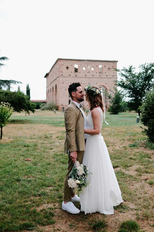 A couple portrait taken outdoors on the grounds of a historic brick building with arched openings and ornate diamond-shaped decorative details, likely a French domaine or abbey. The bride wears a flowing white chiffon gown with a V-neckline and a floral crown of white blooms and greenery, and carries a loose bouquet of white dahlias and eucalyptus. The groom wears a tan/camel suit with a floral boutonniere and white sneakers. The two are facing each other closely, nearly touching foreheads, in a relaxed boho-casual style. The image is a full-length portrait shot with the venue building centered in the soft-focus background.