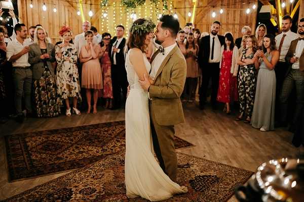 The couple shares their first dance in a rustic barn venue, standing close together on a large vintage-style patterned rug placed on a wooden floor. The bride wears a slim-fit ivory gown with a greenery crown in her hair, while the groom wears an olive-tan tweed suit with a floral or patterned bow tie. Approximately 30–40 guests stand in a circle around them, watching and applauding, dressed in a mix of colorful outfits including floral dresses and formal attire. The barn interior is warmly lit with string Edison bulb lights strung across the wooden ceiling and walls, giving a boho-rustic atmosphere, and the wide-angle shot captures both the dancing couple in the foreground and the full crowd encircling them.
