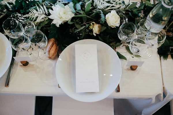 An overhead close-up shot of a wedding reception table setting featuring white round plates, each topped with a slim printed menu card bearing a monogram in a diamond motif. Multiple clear wine and water glasses are arranged at each place setting alongside gold-toned flatware on a white linen tablecloth. Small cork or wooden place card holders are visible at two settings. A lush greenery runner with white roses, peonies, and eucalyptus runs along the center of the table, creating a garden-style tablescape with a clean white and green palette.
