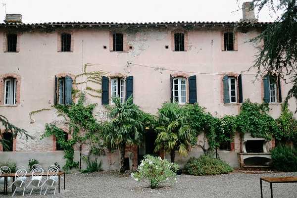 Wide exterior shot of a large French country house with faded pink-terracotta rendered walls, dark teal shutters, and arched brick window surrounds across two stories. The facade is partially covered with climbing vines and features a central palm tree and mature wisteria or ivy trained along the front. A gravel courtyard in the foreground contains a few ornate white metal chairs and a simple wooden bench, along with low shrubs. A built-in stone or brick barbecue structure is visible to the right side of the building. No people are present in the image. Potential venue feature image.
