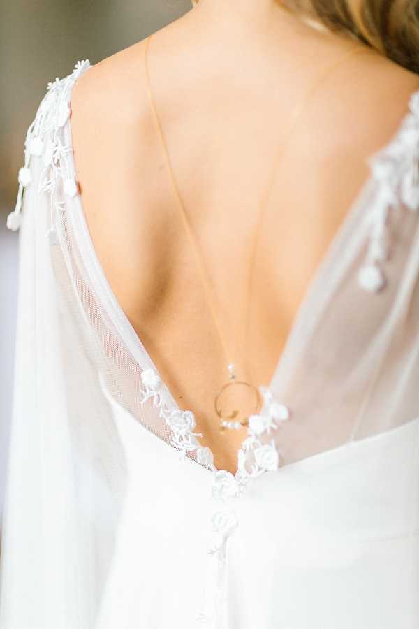Close-up detail shot of a bride's back, showcasing a deep V-back white wedding gown with sheer tulle panels and white 3D floral lace appliqués along the edges and down the back seam. A delicate back-drop necklace with a thin gold chain and small circular pendant rests against the exposed skin. The dress features a clean, modern silhouette with the lace trim providing a romantic decorative accent. The soft, bright natural lighting gives the image a light, airy feel.