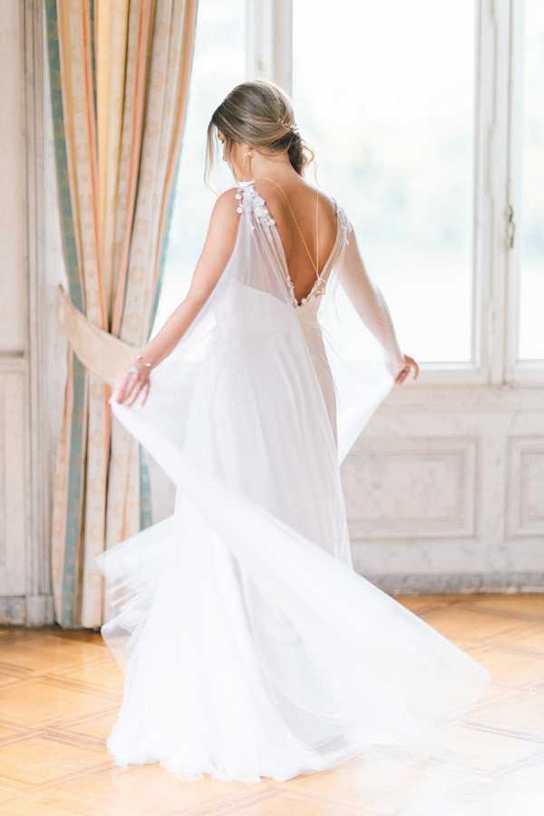 A bride is photographed from behind inside a classic French venue, standing near tall windows with striped peach and teal drapery and ornate white paneled walls. She is wearing a white chiffon gown with a deep open back, delicate floral appliqué details on the shoulders, thin chain back straps, and flowing cape-like sleeves that she is holding out to each side, causing the skirt to swirl. Her dark hair is styled in a low updo with a delicate hair accessory. The image is a full-length portrait shot with soft natural window light creating a bright, airy look, and the parquet wood floor is visible beneath the gown.