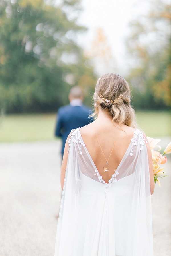 A close-up portrait taken from behind the bride, focusing on the back detail of her white wedding dress, which features a deep V-back, floral appliqué along the shoulder straps, and a flowing chiffon cape overlay. She wears a delicate back necklace with a small pendant and a gold floral hair accessory in her loosely pinned updo. She holds a bouquet with peach and coral blooms visible at the edge of the frame. The groom, dressed in a navy suit, is visible out of focus in the background walking along a gravel path, set against a soft outdoor garden or parkland setting. The overall styling is romantic and boho-influenced, with soft natural light and a muted, airy palette.