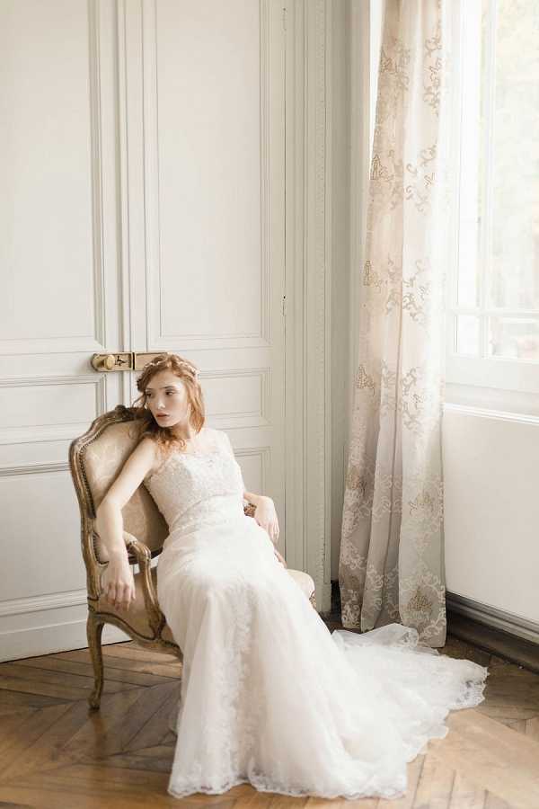 A bridal portrait shot indoors in a classic French interior, featuring a bride seated in a gilded Louis XVI-style armchair with worn upholstery. She wears a fitted ivory lace wedding gown with a flowing train that spreads across the herringbone parquet floor. Her auburn hair is styled up with a delicate floral or braided hair accessory. The room features white boiserie wall paneling and a tall window dressed with an ivory and gold jacquard patterned curtain. Natural window light illuminates the scene from the right. The overall styling is classic and refined, consistent with a Parisian apartment or chateau interior. Medium portrait composition, full-length view.
