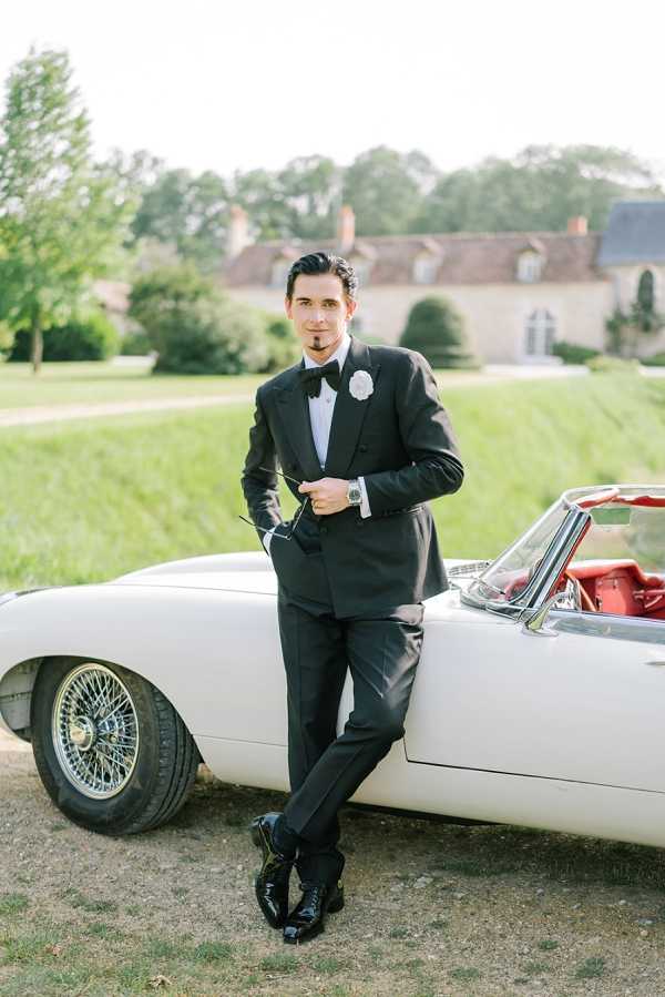 A groom portrait taken outdoors on the grounds of a French chateau, visible in the soft-focus background. The groom leans against a vintage white convertible sports car with red leather interior and wire-spoke wheels, holding a pair of sunglasses in one hand. He wears a black tuxedo with a black bow tie, white dress shirt, a white floral boutonniere, and polished black shoes. The styling is classic black-tie. The image is a medium full-length portrait with a bright, airy exposure.