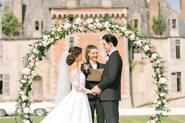 An outdoor wedding ceremony taking place in front of a French chateau, with the couple standing beneath a large circular floral arch decorated with blush pink and white roses, peonies, and lush greenery. The bride wears a full-ballgown with long lace sleeves and a cathedral veil, her hair styled in an updo, while the groom is dressed in a classic black tuxedo; they are holding hands and facing each other. A female officiant stands between them holding a microphone and a small clipboard or booklet. The shot is a medium wide portrait framing all three figures with the chateau's brick and stone facade visible in the background. Potential venue feature image.