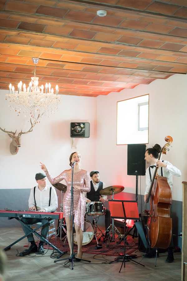 A four-piece live wedding band performs indoors in a room with a terracotta brick ceiling, herringbone wood floor, and white walls. The female vocalist, wearing a short pink sequined dress, sings into a microphone with one arm raised expressively; she is flanked by a keyboard player in a white shirt and suspenders, a drummer in a dark waistcoat, and a double bass player in a white shirt and bow tie. The room is decorated with a crystal chandelier and a mounted stag head on the wall, with warm red ambient uplighting adding to the atmosphere. Wide shot capturing the full band setup, with music stands, a red keyboard, and PA speakers visible.