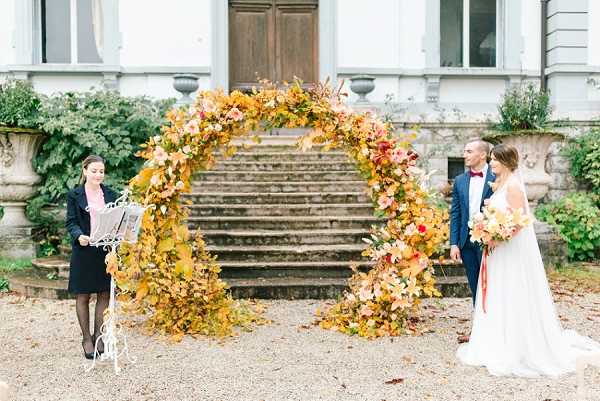An outdoor civil or symbolic ceremony taking place on a gravel courtyard in front of a grand classical French building with stone steps and large ornamental urns. The focal point is a full circular floral arch densely covered in autumn foliage and blooms in shades of amber, burnt orange, gold, coral, and deep red. The couple stands to the right of the arch — the bride wears a white sleeveless gown with a train and holds a bouquet of autumnal orange, red, and peach flowers with a coral ribbon; the groom wears a navy suit with a burgundy bow tie. An officiant stands to the left of the arch, reading from papers on a white ornate lectern. Wide shot capturing the full scene including the building facade as backdrop. The overall palette and styling reflect a rich autumn theme.