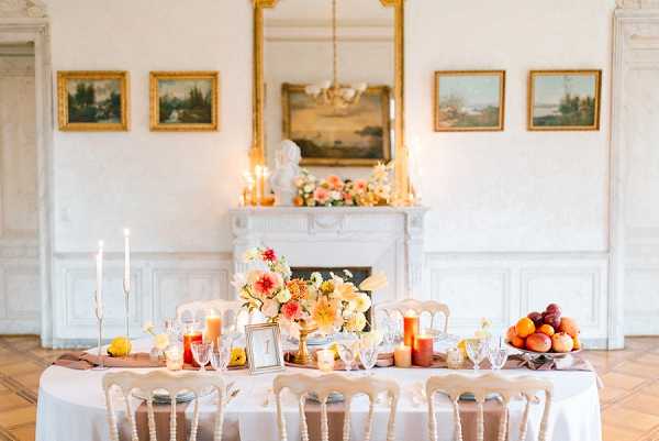 A styled reception tablescape shot in a formal French interior room featuring white paneled walls, an ornate white marble fireplace, a large gilt-framed mirror, and four landscape paintings. The long rectangular table is dressed in a white linen with a terracotta-toned runner and set with crystal glassware, white taper candles in gold holders, and amber-toned pillar candles. The floral centerpiece consists of peach and yellow dahlias, orange blooms, and mixed greenery in a low arrangement, with a matching floral display on the fireplace mantle. A bowl of fresh fruit including peaches and plums sits at one end of the table, and a small framed table number is visible. The chairs are white painted French-style dining chairs. The overall color palette is warm — peach, orange, yellow, and gold tones. Wide interior shot capturing the full table setting and architectural backdrop. Potential venue feature image.