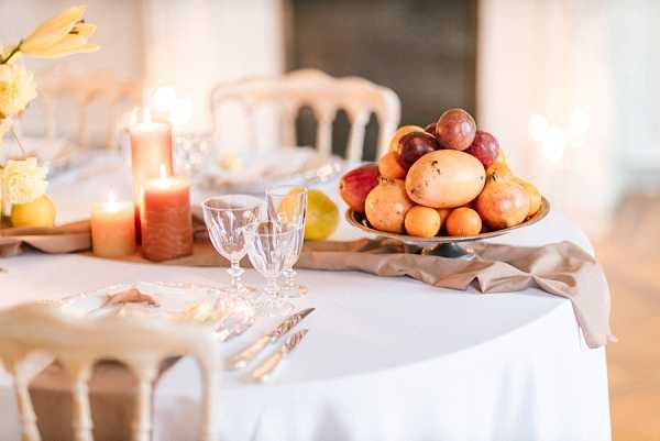 A close-up detail shot of a styled wedding reception tablescape set indoors, likely in a chateau or manor room with a fireplace visible in the background. The table is dressed with a white linen and a draped taupe/greige satin runner, set with crystal glassware, silver flatware, and a metallic pedestal bowl filled with seasonal fruits including peaches, plums, and pears as a centerpiece. Coral and amber pillar candles of varying heights are grouped on a wooden tray alongside yellow tulips or similar blooms, creating a warm, autumnal color palette with a rustic yet refined styling theme. Napoleon-style chairs in a cream finish are visible in the background.