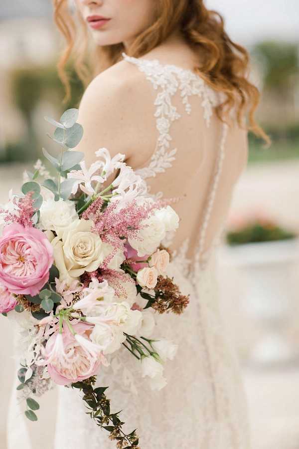 A close-up portrait of a bride shot from behind and slightly to the side, highlighting the back of her ivory lace wedding gown, which features an illusion back panel with delicate floral lace appliqué and a row of fabric-covered buttons running down the spine. She holds a large, loosely arranged bridal bouquet composed of pink garden roses, cream roses, blush peonies, pink astilbe, dusty pink lilies, eucalyptus sprigs, and small bronze-toned filler flowers. The bride has loose, wavy auburn hair worn down. The image is a detail-focused portrait with a shallow depth of field, set outdoors with a softly blurred background.