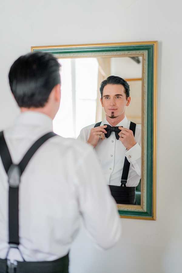A groom getting ready indoors, adjusting a black bow tie while looking at his reflection in a gold and green-framed wall mirror. He is dressed in a white dress shirt with black suspenders and a black cummerbund, with visible cufflinks and a ring on his finger. He has dark slicked-back hair and a small chin beard. The shot is a portrait taken from behind and slightly to the side, with the mirror reflection providing the primary view of his face, against a plain white wall.