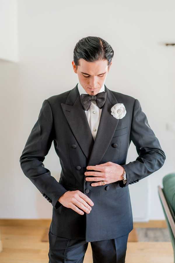 A groom getting ready, photographed indoors against a plain white wall with light wood flooring visible. He is adjusting the buttons of a black double-breasted tuxedo jacket with satin peak lapels, paired with a dark herringbone-patterned bow tie, a white dress shirt with decorative buttons, and a white camellia boutonniere. He wears a gold watch and a gold ring. His dark hair is slicked back. The composition is a mid-length portrait shot with a clean, modern styling aesthetic.