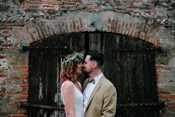 A couple portrait shot outdoors against a rustic brick wall with an arched, weathered dark wooden double door. The bride, wearing a sleeveless white lace dress, has curly auburn hair adorned with a greenery crown featuring small white flowers and eucalyptus-style leaves. The groom wears a tan linen blazer with a floral or patterned bow tie and a white shirt. The two are leaning close together, foreheads nearly touching, smiling. The overall styling is boho-rustic. The image is a medium close-up portrait with a slightly moody, natural light exposure.