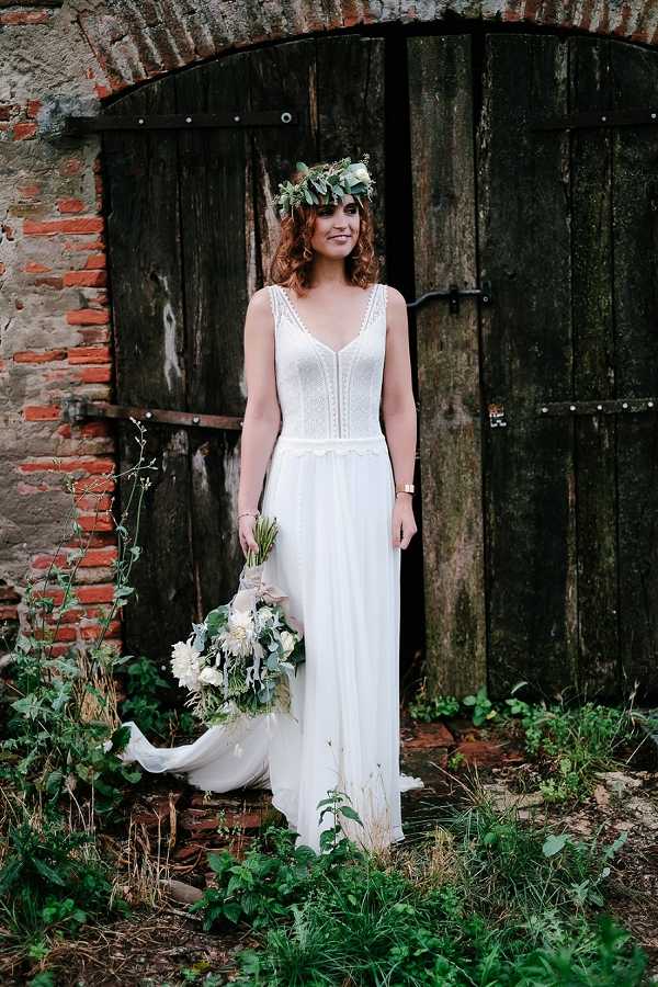 A bridal portrait taken outdoors in front of a rustic weathered wooden double door set within an aged brick and stone archway. The bride stands alone, wearing a floor-length ivory boho-style wedding dress with a lace-detailed bodice, V-neckline, thin straps, and a flowing chiffon skirt with a small train. She wears a lush greenery flower crown with foliage and small blooms, and holds a loose, trailing bouquet featuring white dahlias, dusty blue-grey accents, and eucalyptus with long ribbon streamers in blush and ivory. The styling is distinctly bohemian and rustic. Medium full-length portrait shot.