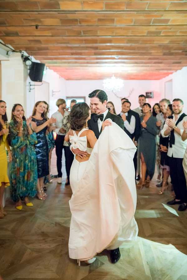 The couple shares their first dance in an indoor reception venue with a warm terracotta tile ceiling, herringbone parquet wood floor, and a chandelier overhead. The groom, wearing a black tuxedo with a black bow tie, lifts the bride slightly off the floor while smiling at her; she wears a white fitted gown with a distinctive large bow detail at the open back. Approximately 20 guests stand in a circle around the dance floor, clapping and watching, dressed in colorful cocktail attire including a bright yellow dress and a teal floral gown. The walls are lit with a warm pink-toned uplighting, and the overall styling is classic and modern. Medium wide shot capturing both the couple and the surrounding guests.