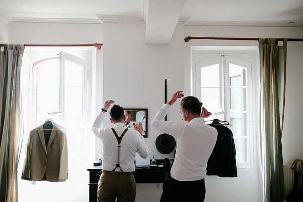 Two men are getting ready in a bright, white-walled interior room, both facing a small wall mirror on a dark dresser and adjusting their hair or accessories. One man is wearing brown suspenders over a white dress shirt with dark trousers, while the other wears a white dress shirt with dark trousers. A light tan/beige suit jacket hangs on a hanger to the left, and a dark jacket is draped to the right near a window. The room features tall French-style windows with neutral linen curtains, suggesting a chateau or manor house setting. The image is a wide shot capturing the getting-ready phase of the wedding day with a relaxed, candid feel.