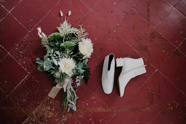 A flat lay detail shot of a bridal bouquet and a pair of white ankle boots placed on deep red terracotta floor tiles. The bouquet features white dahlias, dusty miller, green ferns, dried pampas-style grasses, and eucalyptus foliage, tied with a natural linen ribbon. The white leather ankle boots have a low block heel, side zip, and pearl embellishments at the ankle. The overall styling is modern with bohemian touches, combining structured floral elements with textural dried botanicals.