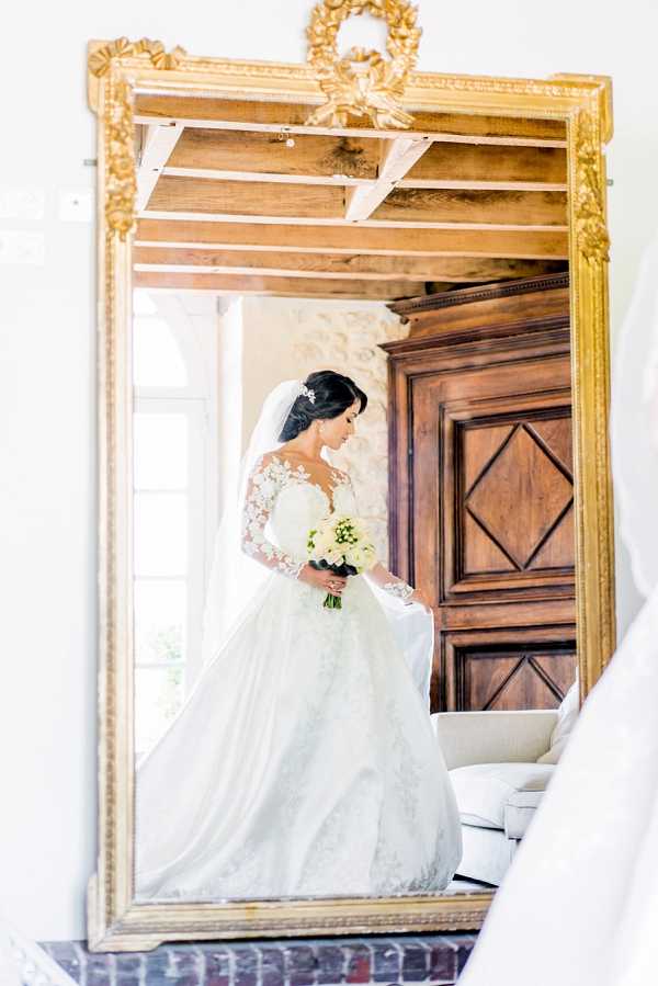 A bridal portrait captured as a reflection in a large ornate gold-framed mirror, shot during the getting-ready phase indoors. The bride wears a full ballgown with sheer lace long sleeves, a cathedral-length veil, and a hair accessory, holding a bouquet of white flowers — likely roses and lily of the valley with green stems. The room features exposed wooden ceiling beams, a large carved dark-wood armoire, and white furnishings, consistent with a classic French chateau interior. The composition is a medium portrait framed within the mirror, with the gold decorative frame — featuring a ribbon-and-wreath carved motif at the top — as the primary foreground element.