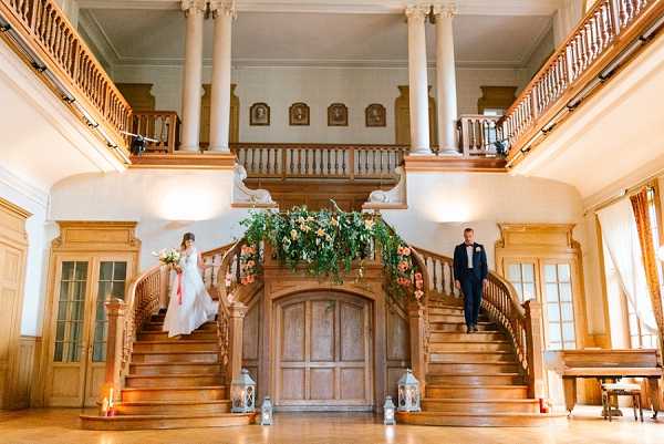 A wide-shot portrait of a bride and groom descending a grand double staircase inside a chateau or manor house, each on opposite sides of the symmetrical wooden staircase. The bride, on the left, wears a full-length white gown and carries a bouquet with coral/pink ribbon trailing down, while the groom, on the right, wears a navy suit with a boutonniere. The central banister between the two staircases is decorated with a lush greenery and cream/yellow floral garland, and several white lanterns are placed at the base of the stairs on the polished wood floor. The interior features warm wood paneling, white columns, a wraparound upper balcony with portrait paintings, and wall sconce lighting giving the space a warm amber tone. The overall decor style is classic and formal. Potential venue feature image.
