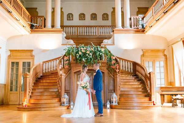 A bride and groom stand facing each other and holding hands in the grand indoor foyer of what appears to be a chateau or historic mansion, positioned in front of a sweeping double wooden staircase with ornate banisters. The bride wears a flowing white gown with a coral-pink ribbon sash and carries a bouquet of peach, coral, and ivory blooms with trailing greenery, while the groom wears a navy blue suit. The staircase landing is decorated with a lush floral arrangement of peach and coral flowers with cascading greenery, and floor-level lanterns with lit candles flank the base of the stairs. The space features warm honey-toned wood, a mezzanine level with portrait frames visible above, and large windows providing bright natural light, giving the interior a classic, formal aesthetic. Wide portrait shot capturing the full architectural setting. Potential venue feature image.