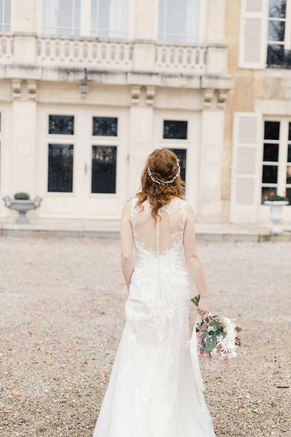 A bridal portrait taken outdoors on a gravel courtyard in front of a classic French chateau facade with tall shuttered windows, stone balustrades, and decorative urns. The bride is photographed from behind, facing the building, wearing a fitted ivory lace gown with an illusion back detail, a row of covered buttons running down the spine, and a modest train. Her auburn hair is styled in a loose updo adorned with a delicate crystal or pearl hair vine. She holds a loose, garden-style bouquet featuring white blooms, blush pink flowers, and eucalyptus foliage at her side. The composition is a mid-length portrait shot with the chateau softly blurred in the background. Potential venue feature image.