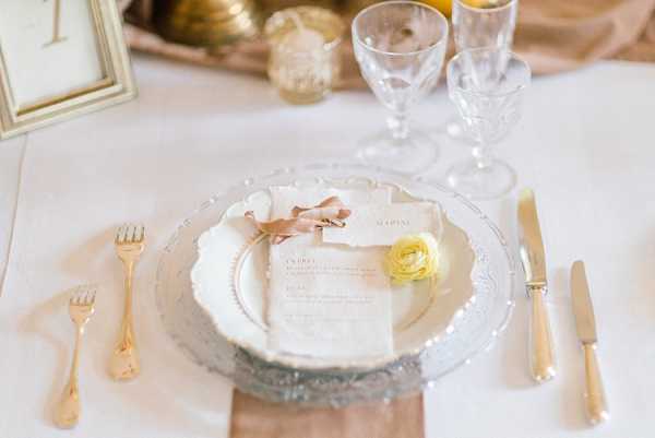 A close-up detail shot of a wedding reception place setting on a white linen tablecloth. The setting features a silver ornate charger plate layered with a white dinner plate, on top of which sits a cream-colored menu card tied with a dusty rose silk ribbon and accented with a single yellow ranunculus bloom. Gold flatware flanks the plates, and a folded blush linen napkin is visible beneath the charger. Crystal cut-glass goblets, gold-rimmed glassware, and a gold-framed table number card are visible in the soft-focus background alongside small candle votives. The overall decor palette combines white, gold, blush, and soft yellow in a classic, refined style.