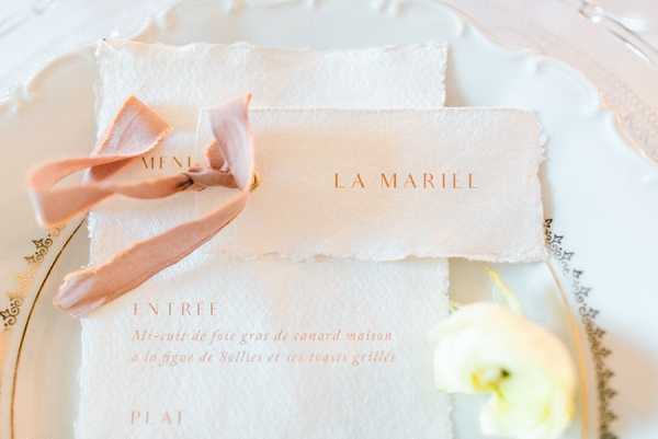 A close-up flat lay of French wedding stationery displayed on a white charger plate with gold rim detailing. The stationery consists of deckle-edged handmade paper pieces, including a menu card printed in rose gold/copper typography and a place card reading 'LA MARIÉE' (the bride). A soft blush pink silk ribbon is loosely tied around the stationery. A small white ranunculus bloom is partially visible at the bottom right corner. The overall palette is ivory, blush, and rose gold, suggesting a refined classic French wedding aesthetic.