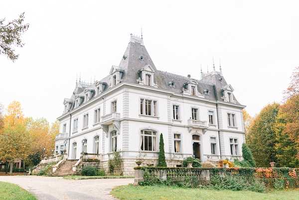 Wide-angle exterior shot of a French château featuring classic 19th-century architecture with a steep slate mansard roof, dormer windows, ornate stone facades, and a grand stone staircase leading to the entrance. The building is white/cream rendered with symmetrical window arrangements and decorative balustrades along the perimeter. No people or wedding party are visible in the frame. Potential venue feature image.